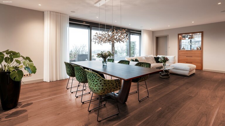 Dunkler mafi Naturholzboden Nuss in einem großen Wohn- Essbereich mit Esstisch, Stühlen, Sofa und Vitrine. Mittig dahinter Fenster und Türen welche auf eine Terrasse führen